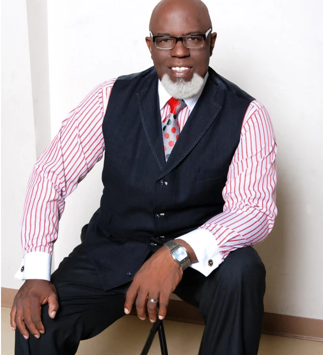 Minister James wearing black vest and pants with red and white striped shirt sitting in studio